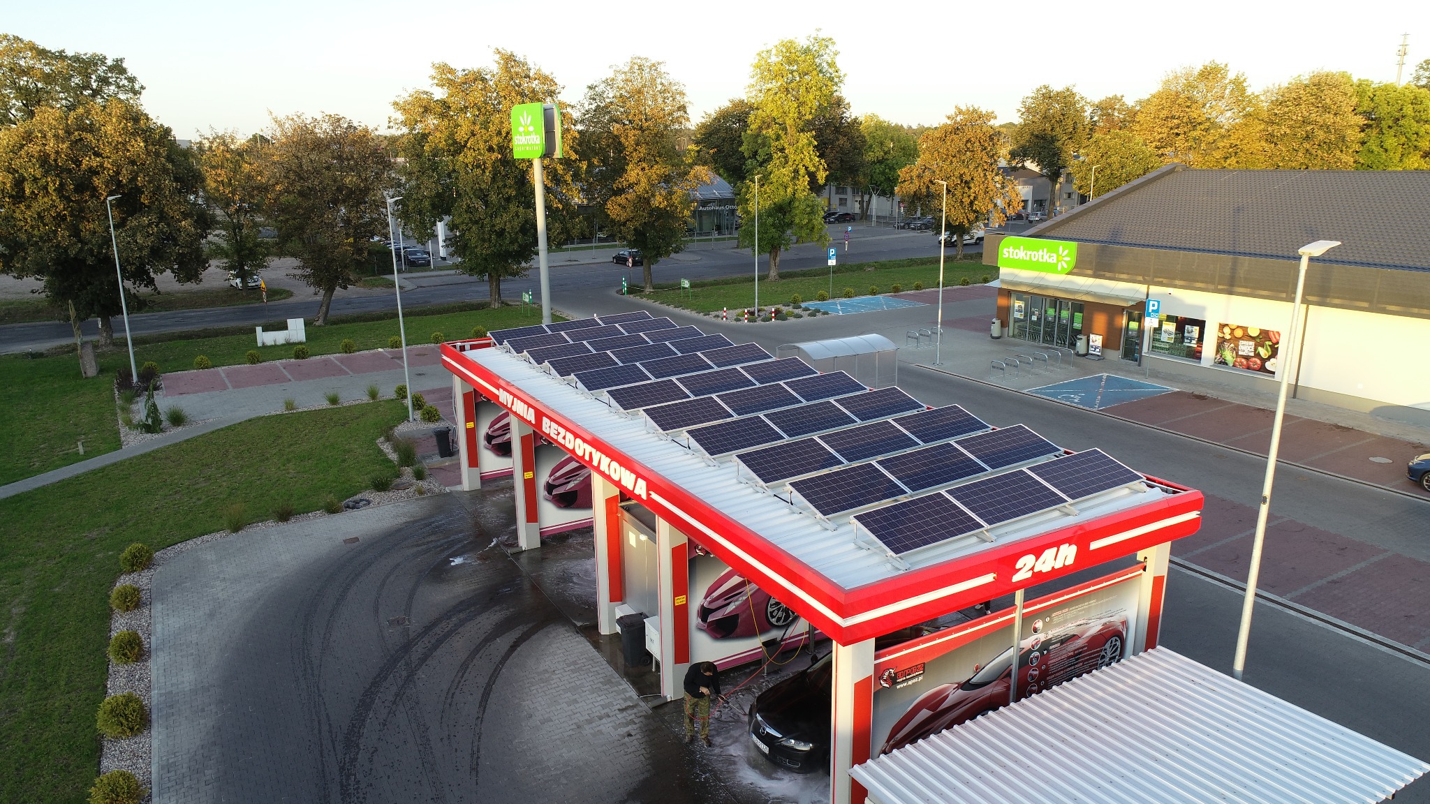 Widok z góry na bezdotykową myjnię samochodową z panelami fotowoltaicznymi na dachu, obok sklep Stokrotka, w tle drzewa.