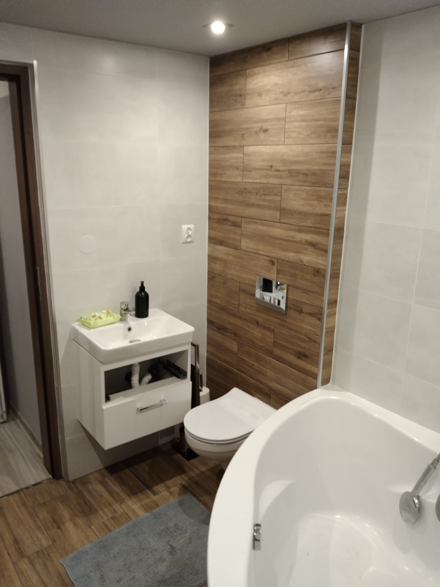 Nowoczesna łazienka z drewnopodobnymi płytkami na ścianie, białą armaturą i szarą wycieraczką.