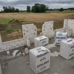 Wznoszone ściany domu z białych bloczków Solbet, kilka opakowań bloczków na mokrej posadzce, widok na pole i pochmurne niebo.