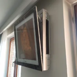 Klimatyzator LG Artcool Gallery z obrazem Klimta w folii ochronnej, zamontowany na ścianie w jasnym wnętrzu, widoczne okno z firanką.