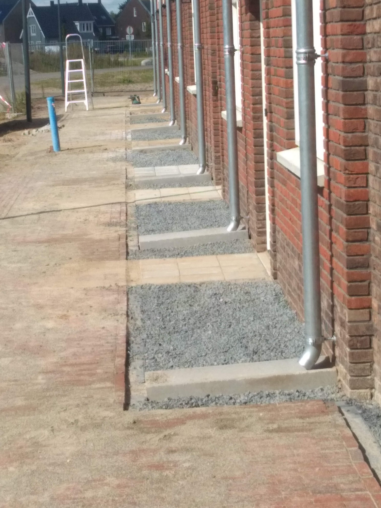 Rząd budynków z cegły z zamontowanymi rynnami spustowymi, obok wyłożony chodnik z kostki i żwiru, widoczne prace wykończeniowe.