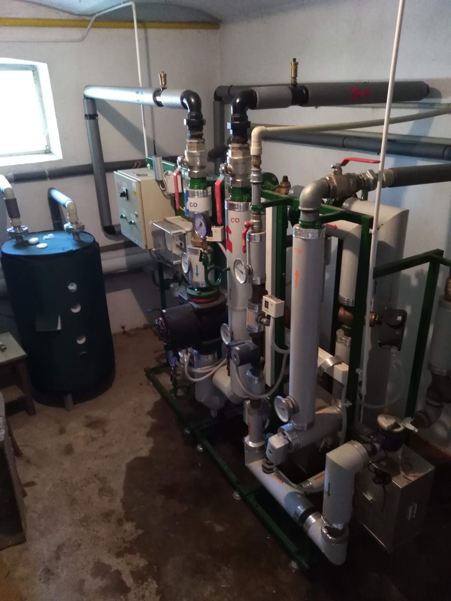Złożona instalacja hydrauliczna z rurami, zaworami, manometrami i zbiornikiem akumulacyjnym w pomieszczeniu technicznym. Widoczne oznaczenia 'CO' i 'CW' na rurach.