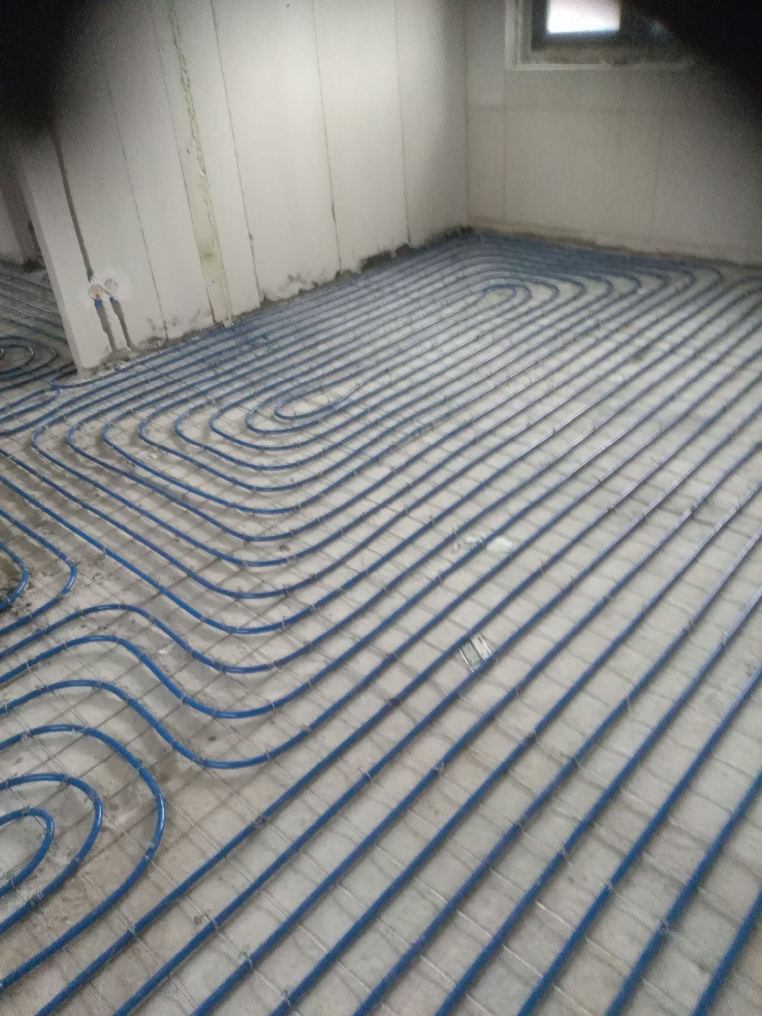 Niebieskie rury ogrzewania podłogowego ułożone w spiralny wzór na siatce w surowym pomieszczeniu, widoczne przyłącza do ściany.