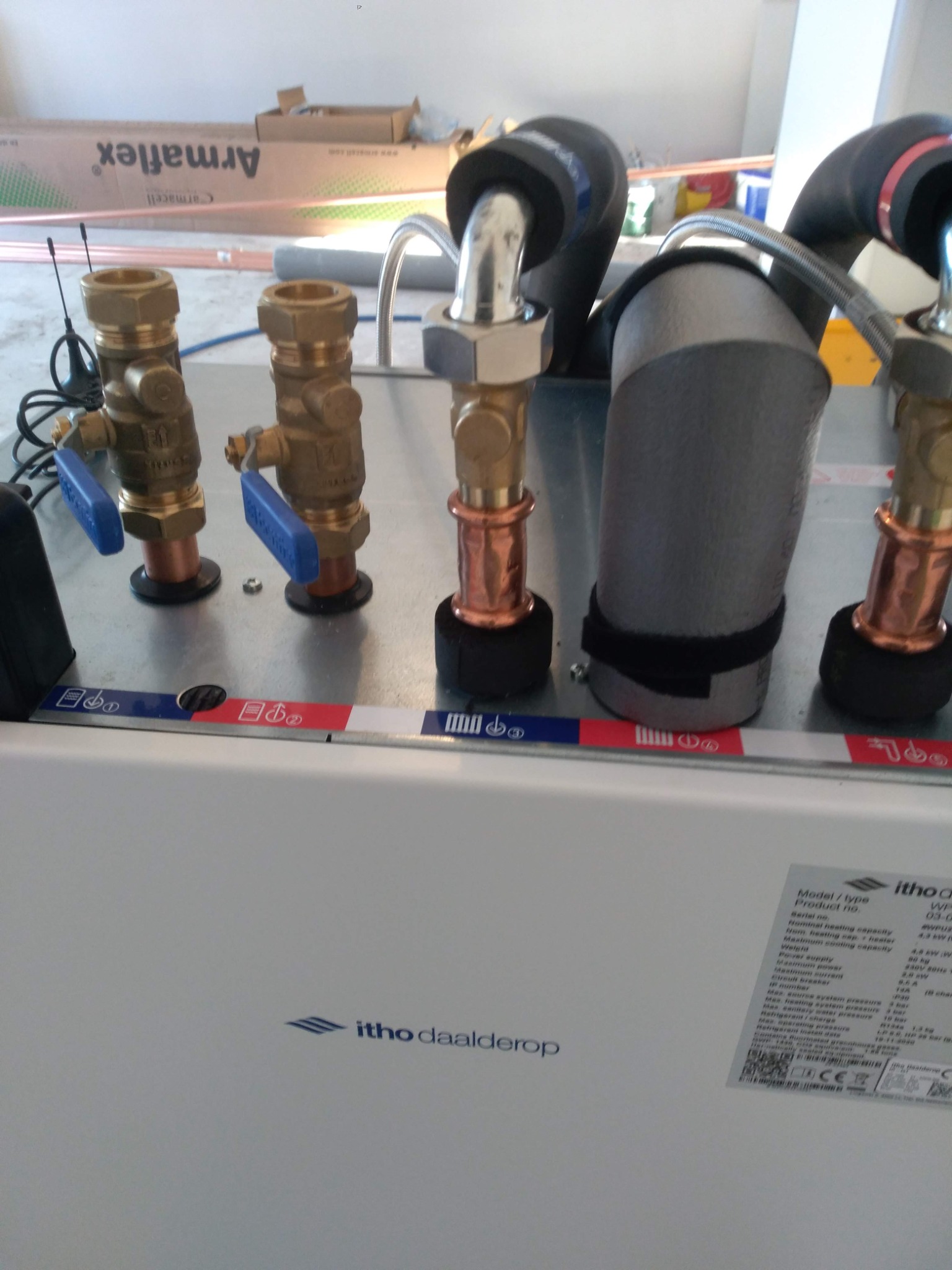 Szczegółowe ujęcie instalacji hydraulicznej z mosiężnymi zaworami z niebieskimi uchwytami, rurami miedzianymi i izolowanymi przewodami, zamontowanej na białej obudowie urządzenia Itho Daalderop,...