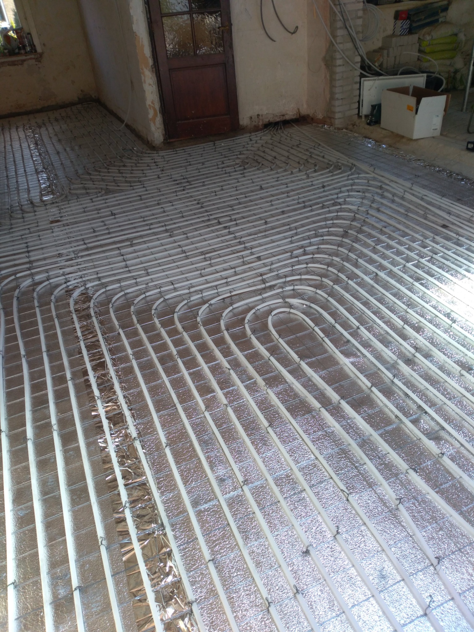 Rozłożona instalacja ogrzewania podłogowego z rur na folii izolacyjnej, widoczne metalowe zaczepy mocujące rury do siatki, surowy stan wykończenia wnętrza.