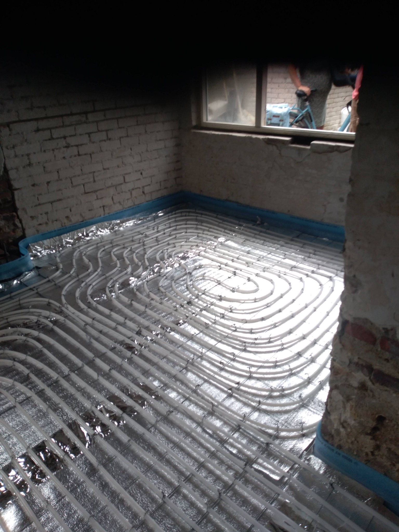 Montaż rur ogrzewania podłogowego na folii aluminiowej w surowym pomieszczeniu z ceglanymi ścianami i widocznym oknem.