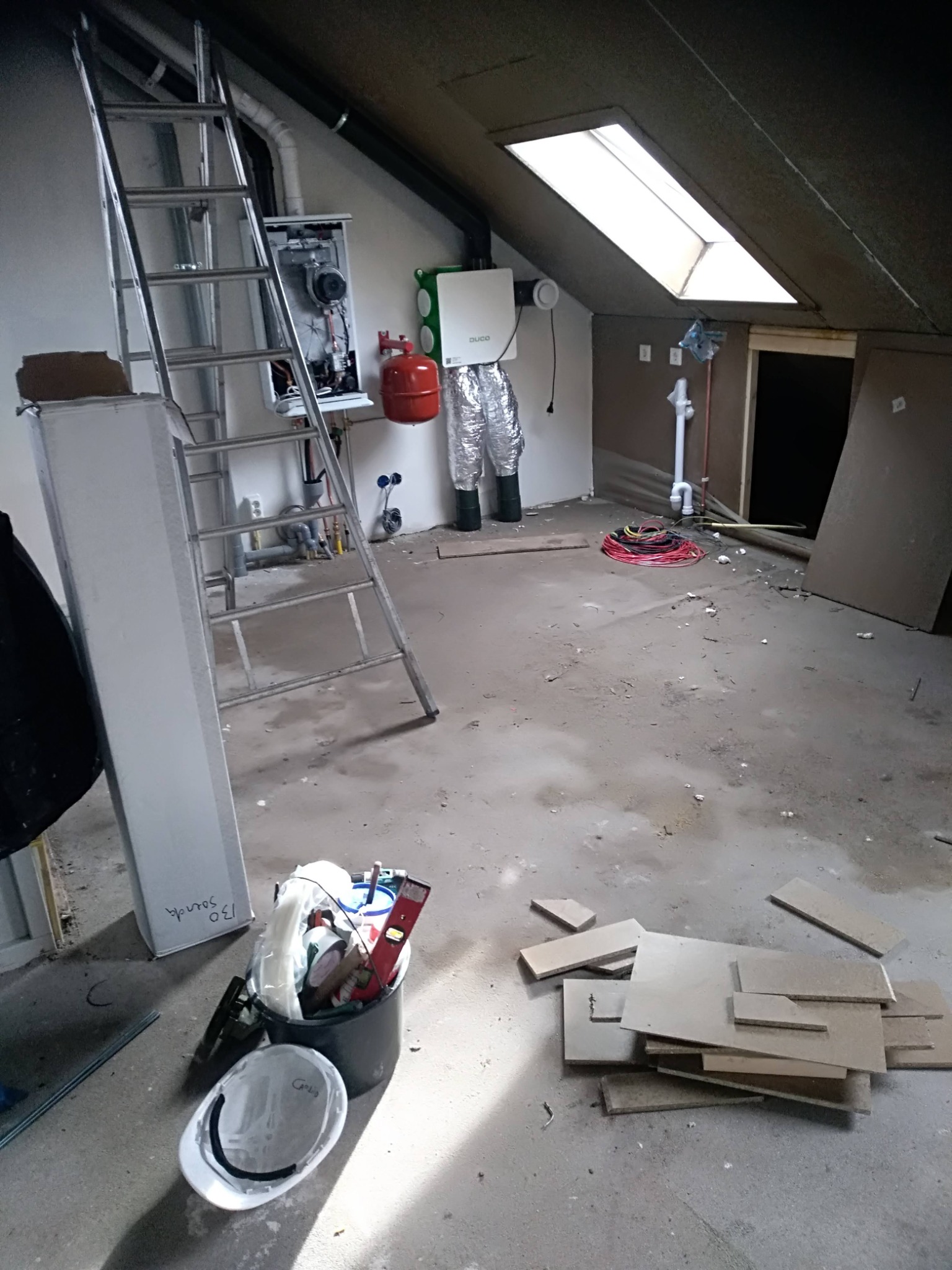 Poddasze w Zeewolde w trakcie prac instalacyjnych: widoczny piec, elementy wentylacji, przewody, drabina i narzędzia w wiadrze. Stan surowy, przygotowanie do dalszych prac.