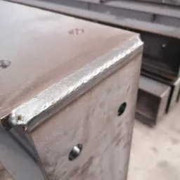 Metalowy profil o przekroju prostokątnym, z widocznymi spawami na krawędziach i otworami, ustawiony pionowo; w tle inne podobne elementy.