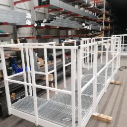 Biała, metalowa konstrukcja balustrady z kratownicą podłogową, tymczasowo podparta drewnianymi klockami, w hali produkcyjnej z regałami magazynowymi w tle.