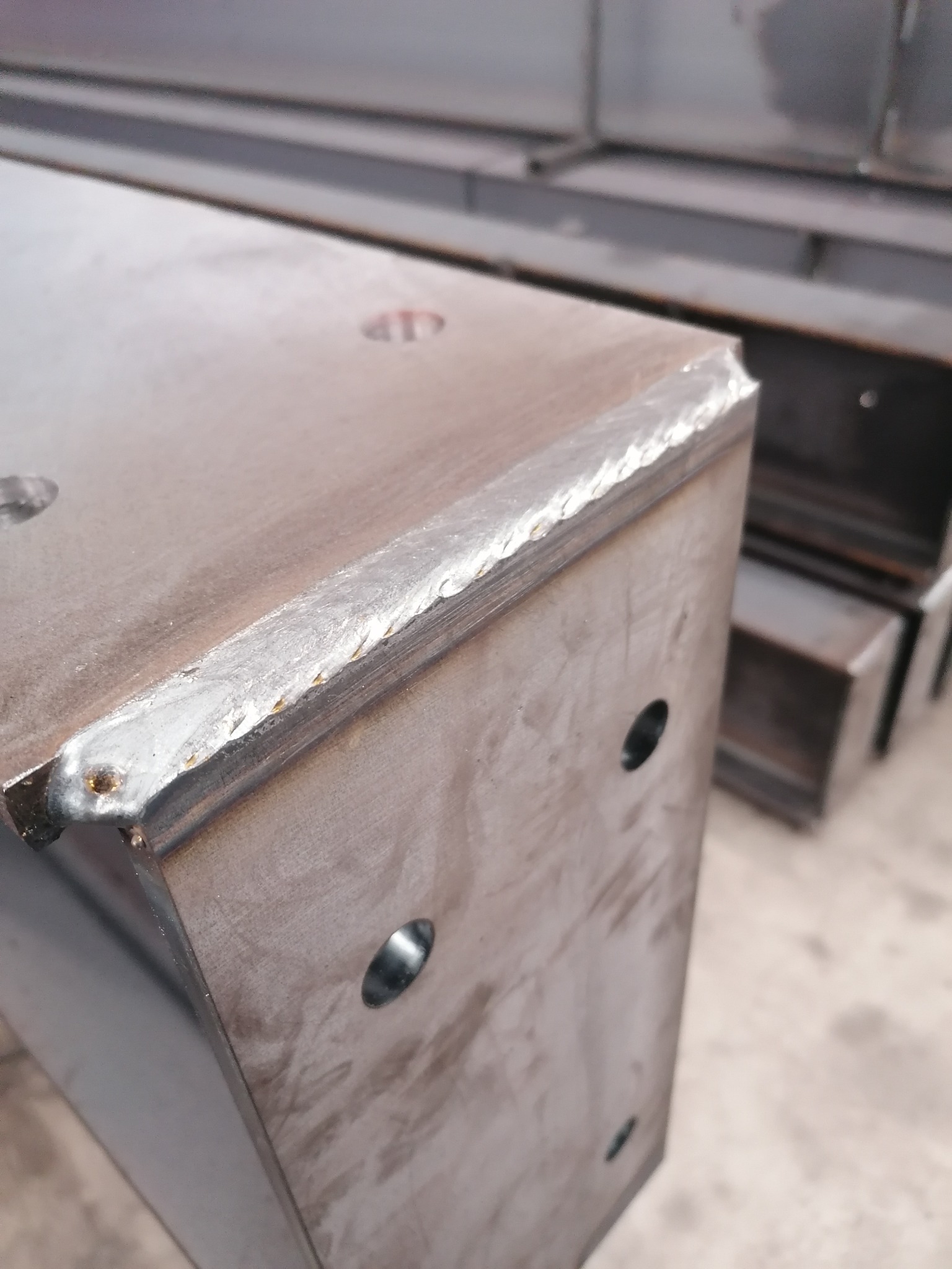 Metalowy profil o przekroju prostokątnym, z widocznymi spawami na krawędziach i otworami, ustawiony pionowo; w tle inne podobne elementy.