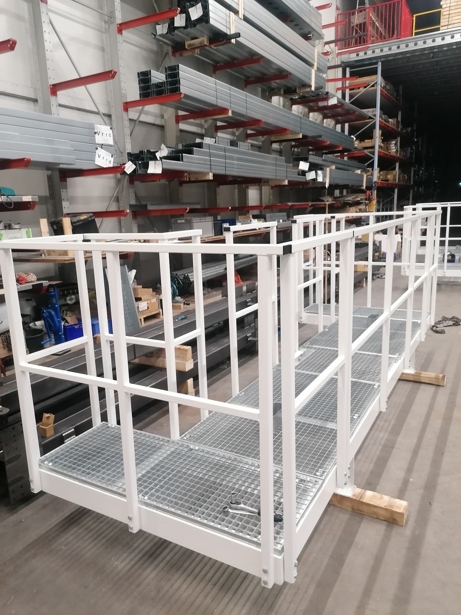 Biała, metalowa konstrukcja balustrady z kratownicą podłogową, tymczasowo podparta drewnianymi klockami, w hali produkcyjnej z regałami magazynowymi w tle.