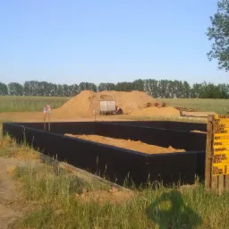 Przygotowany, czarny szalunek fundamentów na piaszczystym podłożu z widoczną tablicą informacyjną budowy i stertą piasku w tle.