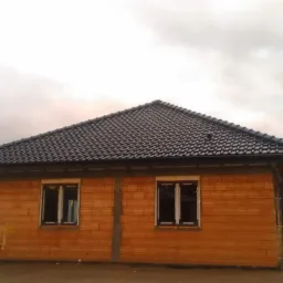 Surowy stan budynku jednorodzinnego z cegły ceramicznej, z zamontowanymi oknami i ciemną dachówką, widoczne rynny i fragment krajobrazu w pochmurny dzień.