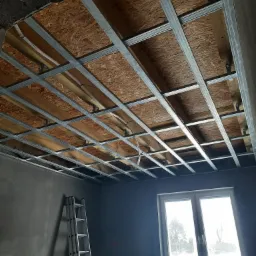 Stelaż z profili metalowych zamontowany do sufitu z płyt OSB, przygotowany pod montaż okładziny, z widoczną drabiną opartą o szarą ścianę i oknem w tle.