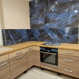 Nowoczesna kuchnia z drewnianymi szafkami, blatem w kolorze jasnego drewna i okładziną ściany imitującą marmur w odcieniach granatu i błękitu, z widoczną płytą gazową i oknem z żaluzjami.