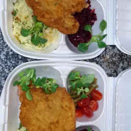 Dwa zestawy obiadowe w białych, plastikowych opakowaniach: panierowany kotlet, ziemniaki z koperkiem, sałatka z buraczków i mieszanka warzyw z pomidorkami cherry, udekorowane świeżymi listkami.