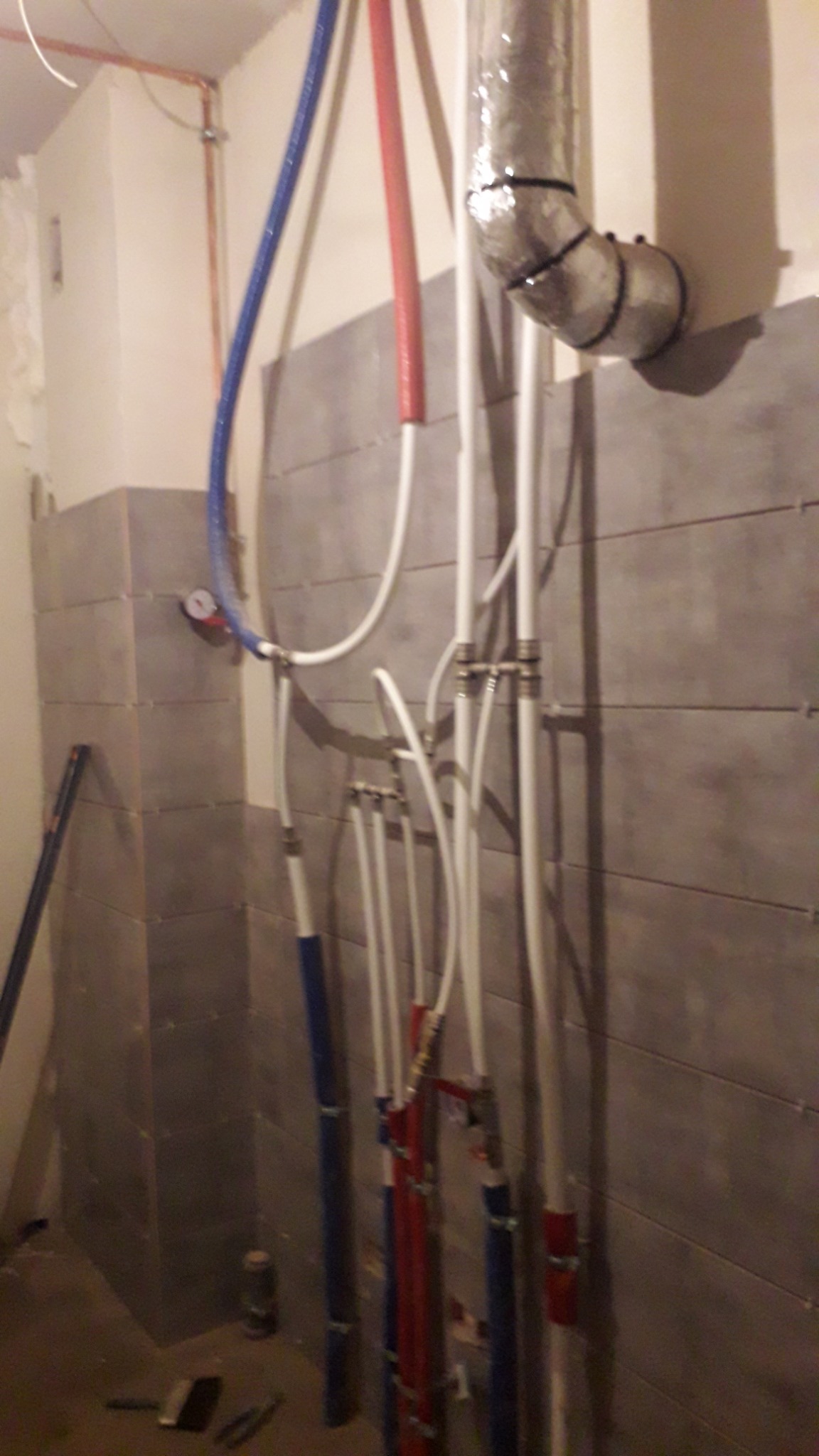 Szare płytki na ścianie w trakcie remontu łazienki, widoczne rury instalacji wodnej (czerwone i niebieskie) oraz fragment wentylacji.