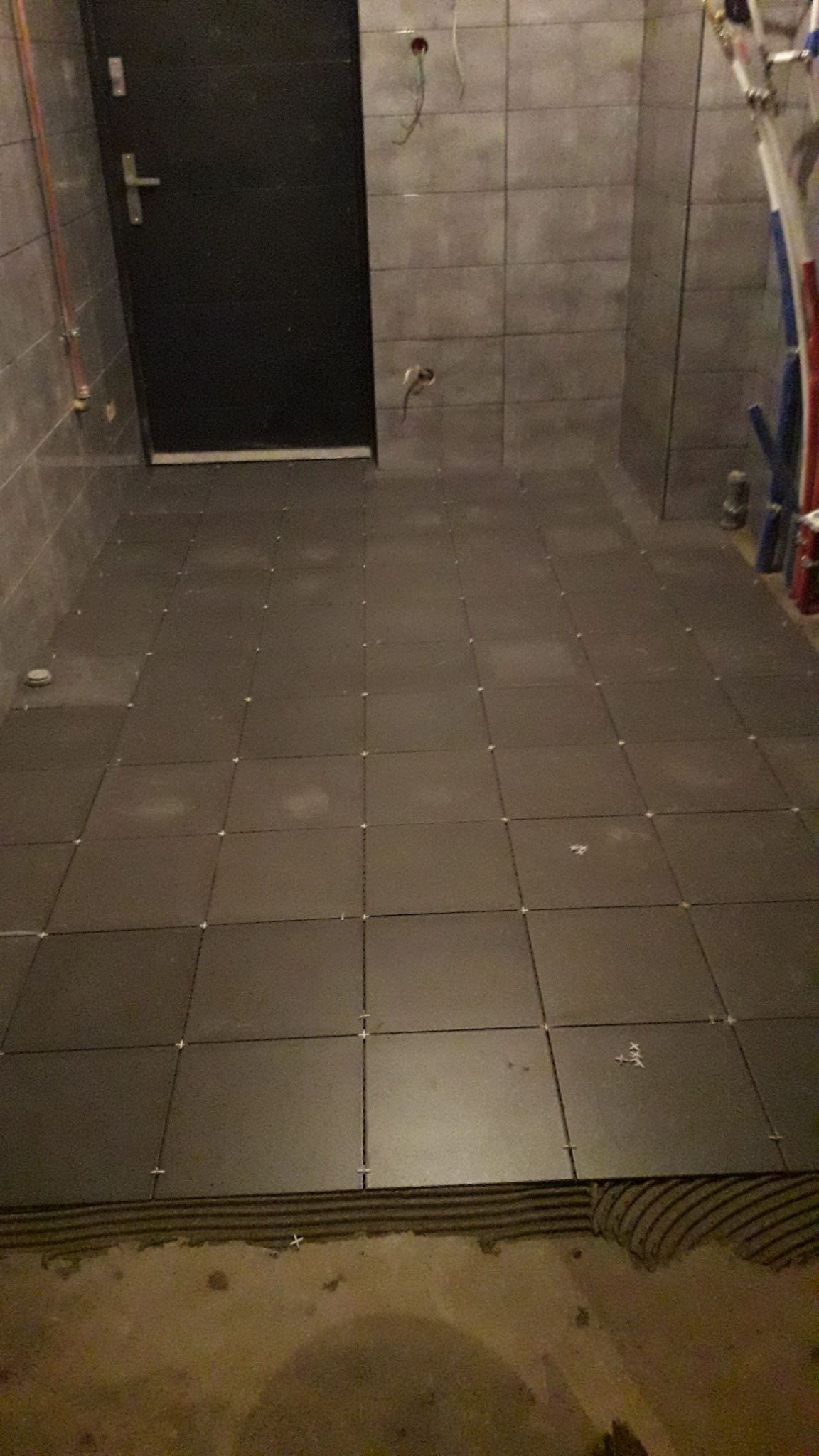 Układanie ciemnoszarych płytek podłogowych w łazience z widocznymi krzyżykami dystansowymi i fragmentem ściany z instalacją hydrauliczną.