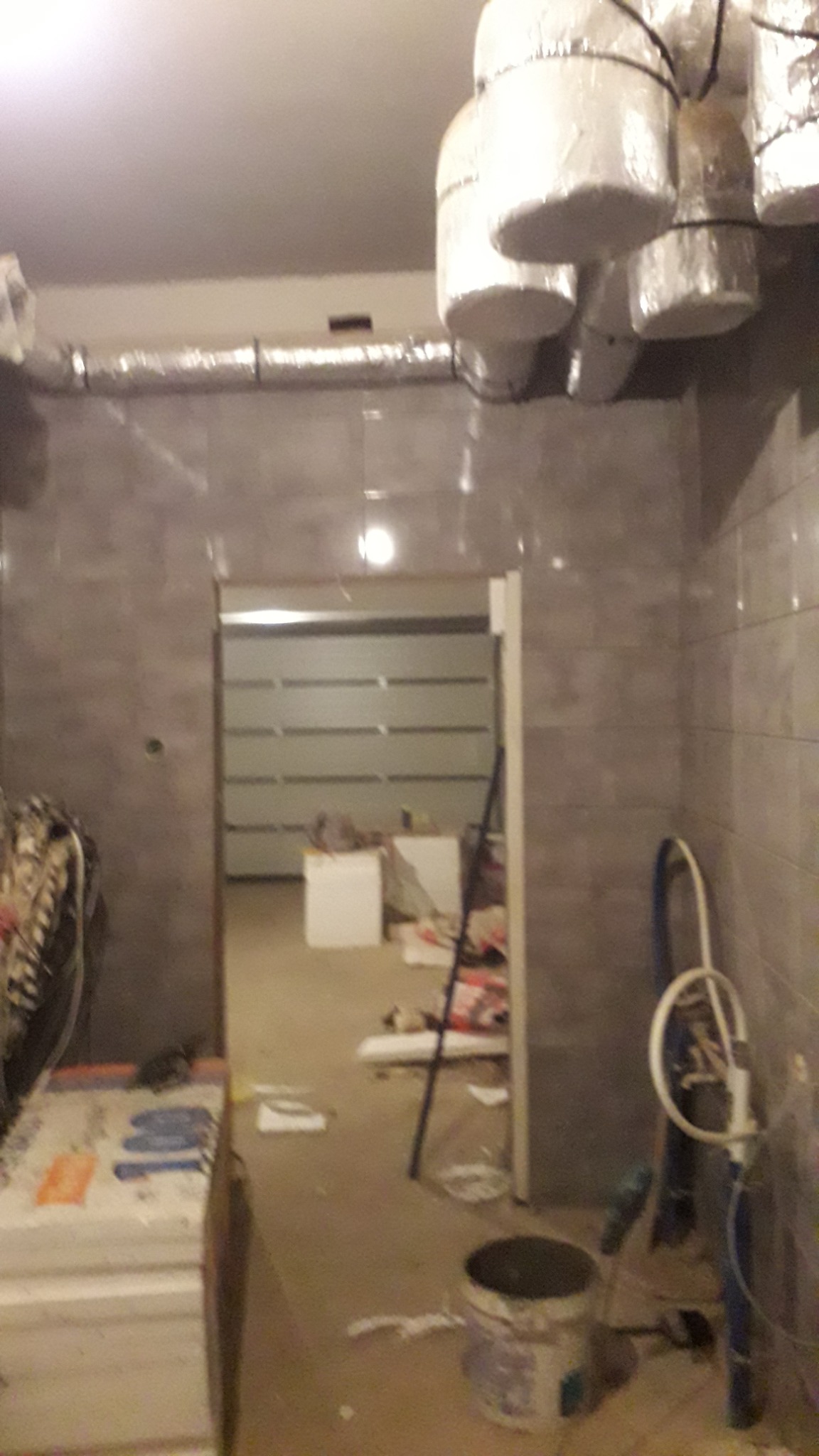 Remont łazienki w trakcie: widoczne szare płytki na ścianach, zabezpieczone folią lampy sufitowe, rury, wiadro z zaprawą i materiały budowlane w pomieszczeniu z otwartym przejściem do innego pokoju.