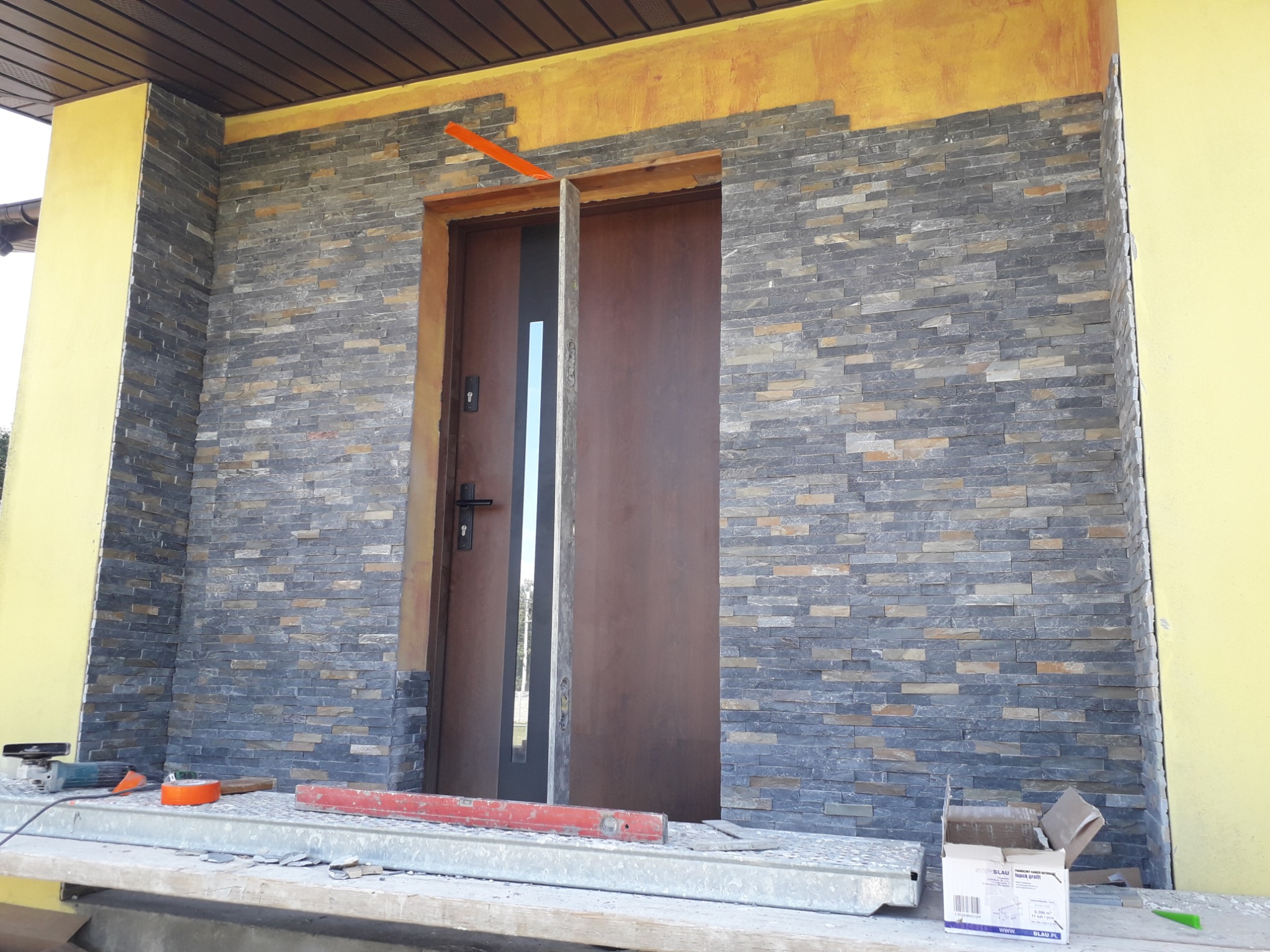 Montaż dekoracyjnej okładziny kamiennej na elewacji wokół wejścia, widoczne narzędzia i poziomica, częściowo wykończona ściana z szarego kamienia łupkowego.