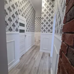 Korytarz po remoncie z tapetowanymi ścianami w biało-szare wzory, białą boazerią, panelami podłogowymi i fragmentem ściany z czerwonej cegły.