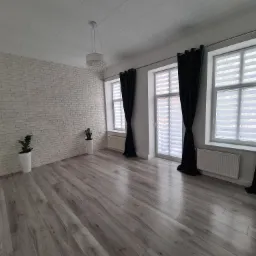 Jasne, puste wnętrze po remoncie z szarą podłogą imitującą drewno, białą ceglaną ścianą, trzema oknami z roletami i ciemnozielonymi zasłonami oraz roślinami w białych donicach.