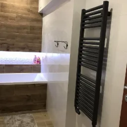 Nowoczesna łazienka z grzejnikiem drabinkowym w kolorze antracytowym, drewnopodobne płytki na ścianie, biała wanna z podświetleniem LED i szary dywanik.