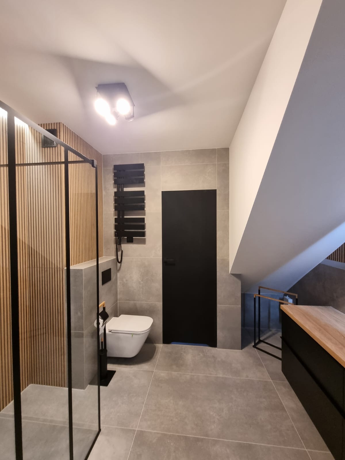 Nowoczesna łazienka z kabiną prysznicową typu walk-in z czarnymi ramami, grzejnikiem drabinkowym w kolorze czarnym, toaletą i czarnymi drzwiami, wykończona szarą płytką i drewnianą okładziną.