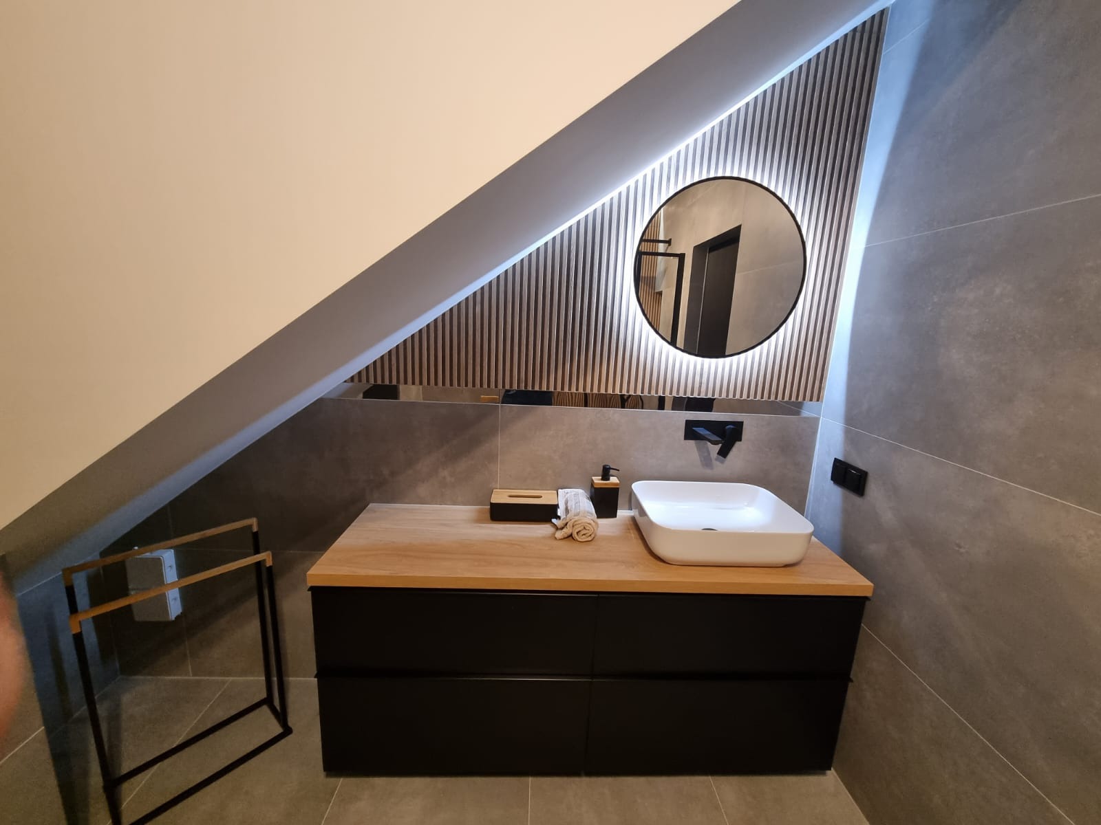 Nowoczesna łazienka na poddaszu z okrągłym lustrem podświetlanym LED, drewnianą szafką z czarnymi szufladami i białą umywalką nablatową.