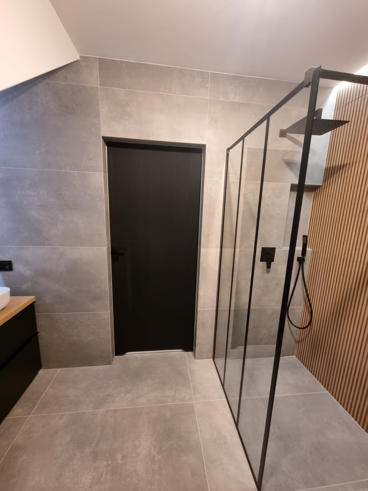 Nowoczesna łazienka z czarną kabiną prysznicową typu walk-in, szarymi płytkami i drewnianą ścianą dekoracyjną z lamelami.