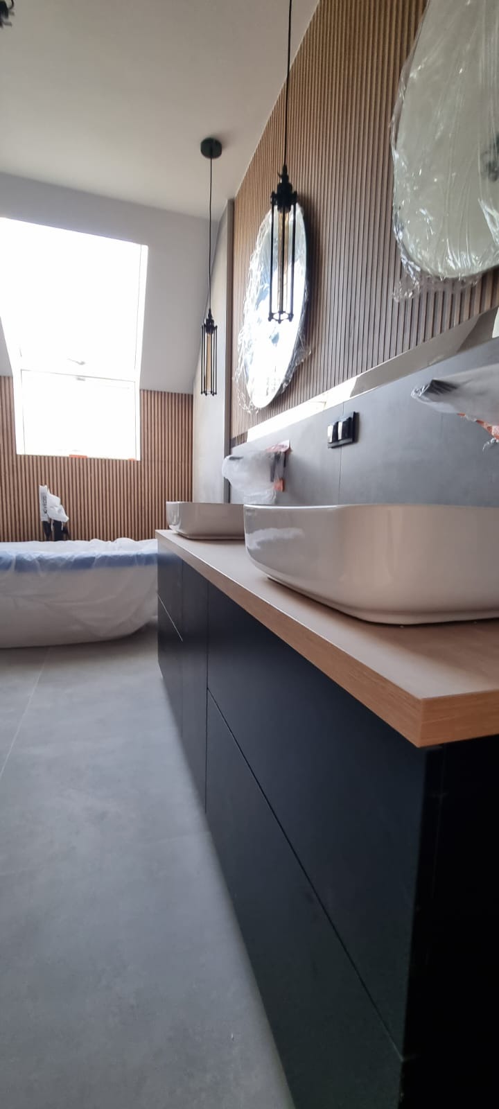 Nowoczesna łazienka w trakcie wykańczania: dwie białe umywalki nablatowe, czarne szafki pod umywalkami z blatem imitującym drewno, pionowe panele ścienne z drewna, okrągłe lustra w folii ochronnej,...