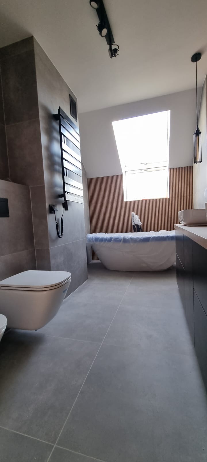 Nowoczesna łazienka w szarościach z drewnianymi panelami ściennymi, podwieszana toaleta, grzejnik drabinkowy i wanna zabezpieczona folią na czas prac wykończeniowych.