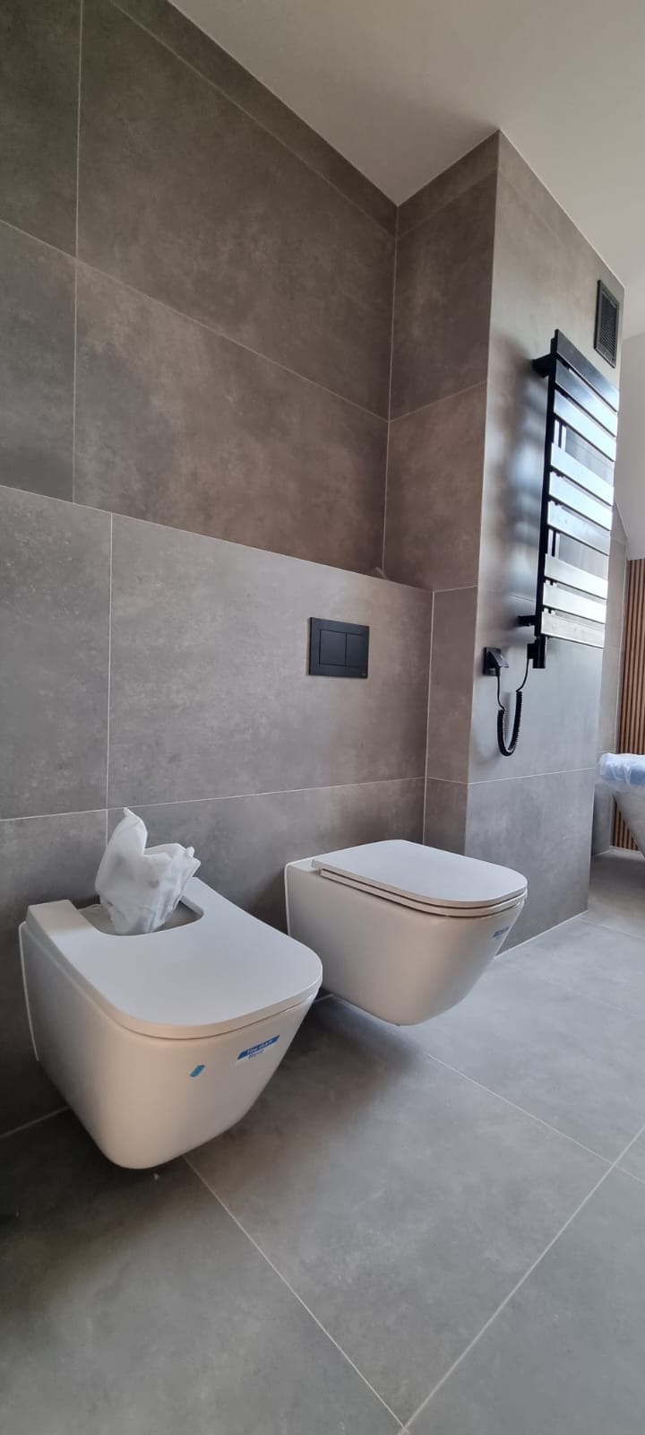 Nowoczesna łazienka z szarymi płytkami, wiszącą toaletą i bidetem z zabezpieczeniem folią, grzejnik drabinkowy na ścianie.