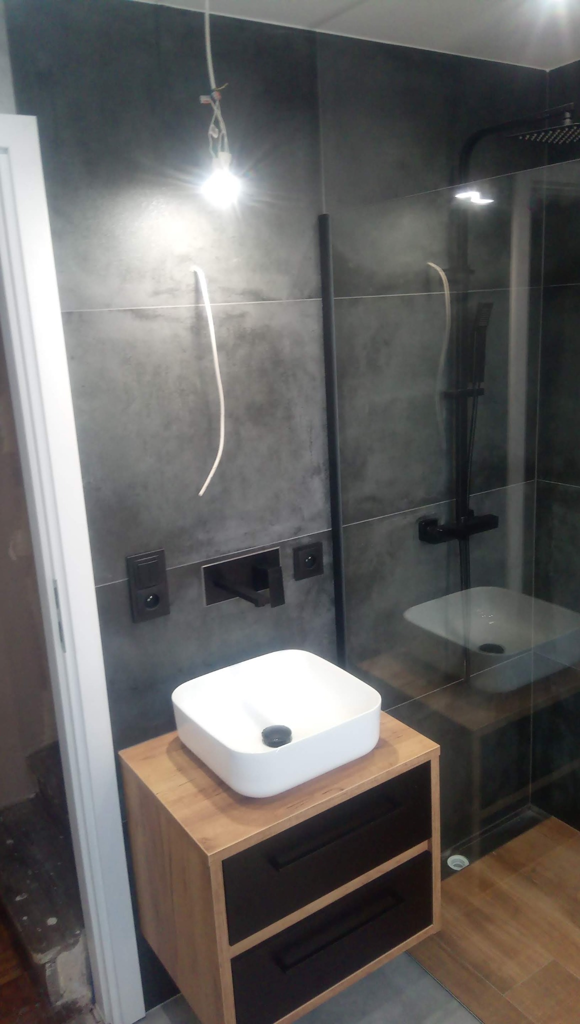 Nowoczesna łazienka w trakcie wykańczania z ciemnoszarymi płytkami, białą umywalką nablatową na drewnianej szafce z czarnymi szufladami, czarną armaturą i kabiną prysznicową z czarnym panelem.