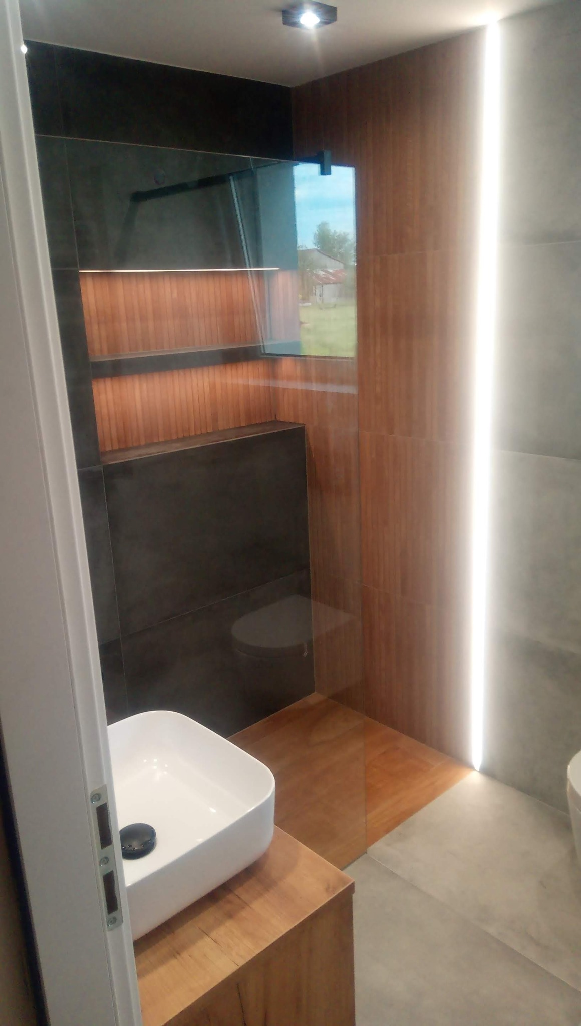 Nowoczesna łazienka z drewnianymi i szarymi płytkami, podświetlaną wnęką prysznicową i białą umywalką nablatową na drewnianym blacie.