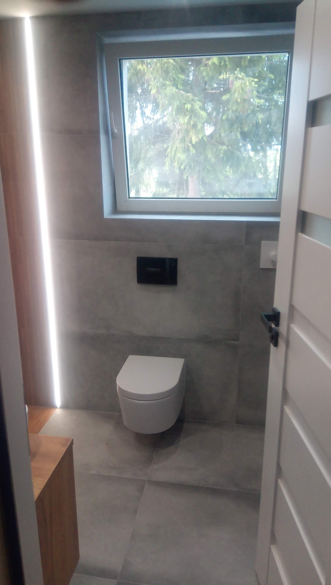 Nowoczesna toaleta podwieszana w szarej łazience z oknem, widok przez otwarte drzwi, oświetlenie LED w pionie, minimalistyczny styl.