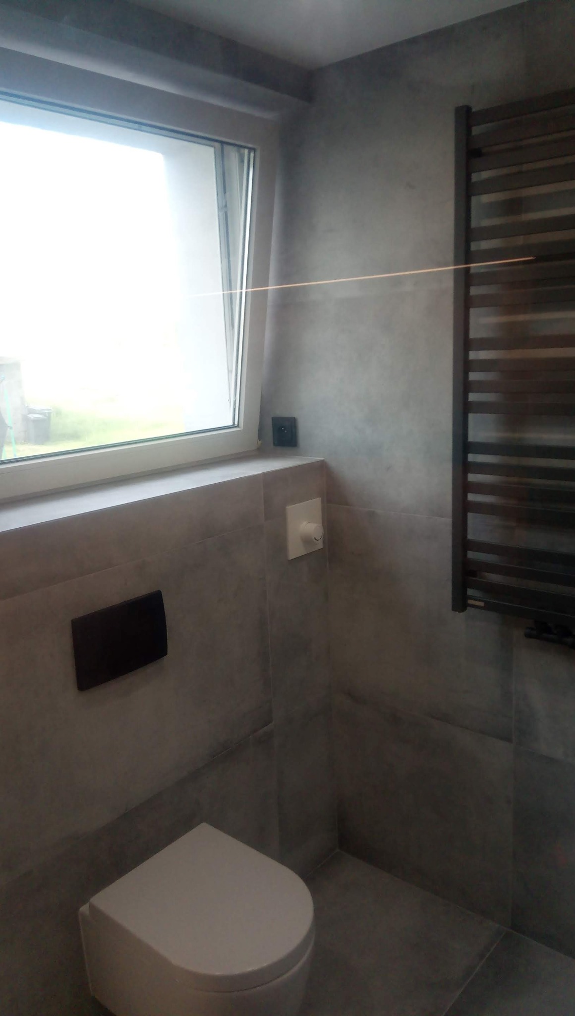 Wykończona łazienka w Debrznie z szarymi płytkami, wiszącą toaletą i grzejnikiem drabinkowym, widok z niskiego kąta.