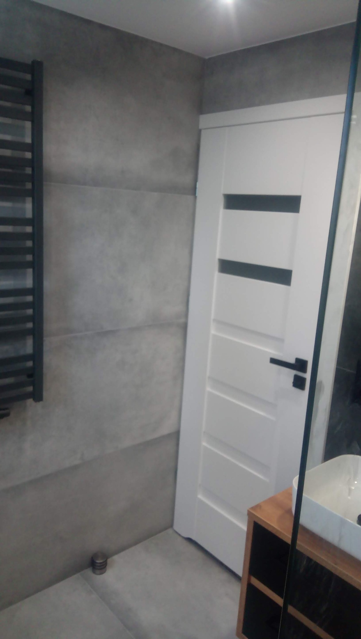 Białe drzwi wewnętrzne z czarną klamką i przeszkleniami, widoczne na tle szarej ściany z grzejnikiem drabinkowym w łazience po remoncie.