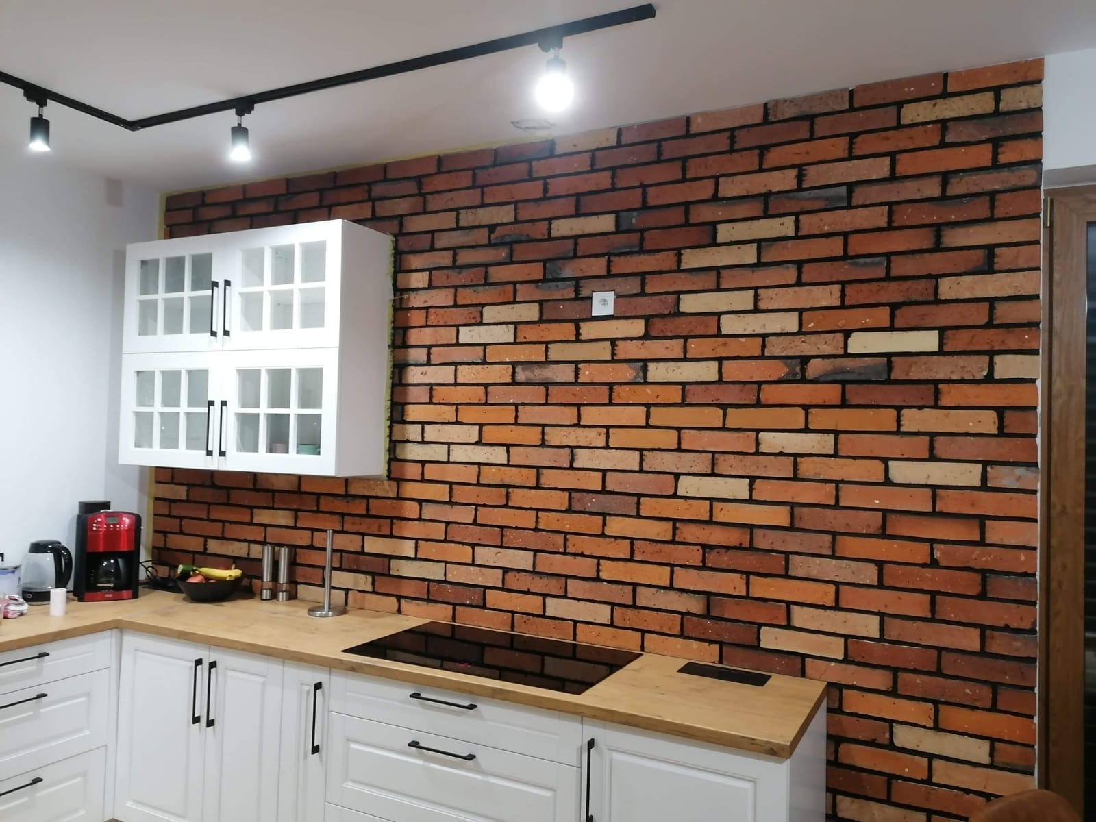 Kuchnia z ceglaną ścianą, białymi szafkami z przeszklonymi frontami, drewnianym blatem i czarnymi uchwytami, oświetlona szynoprzewodem z reflektorami.