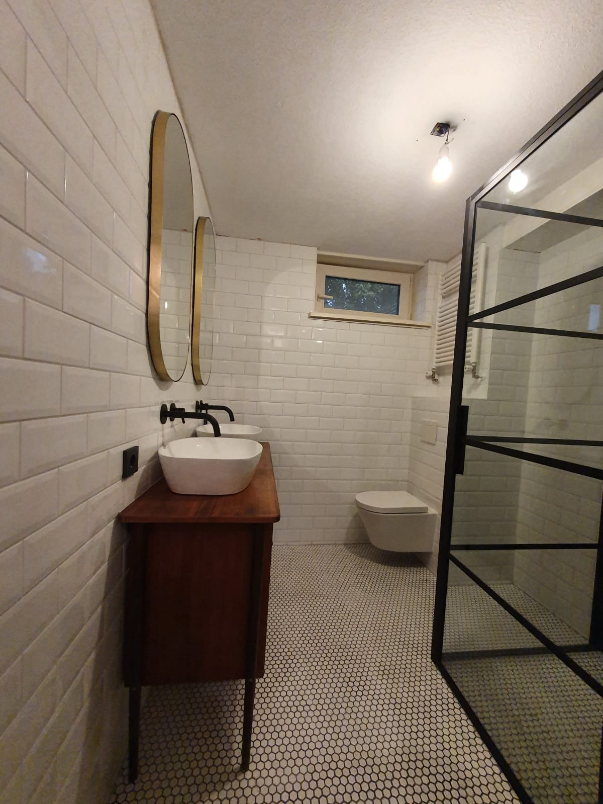 Nowoczesna łazienka z białymi płytkami metra, okrągłymi lustrami w złotych ramach, umywalką nablatową na drewnianej szafce i kabiną prysznicową z czarnymi profilami.