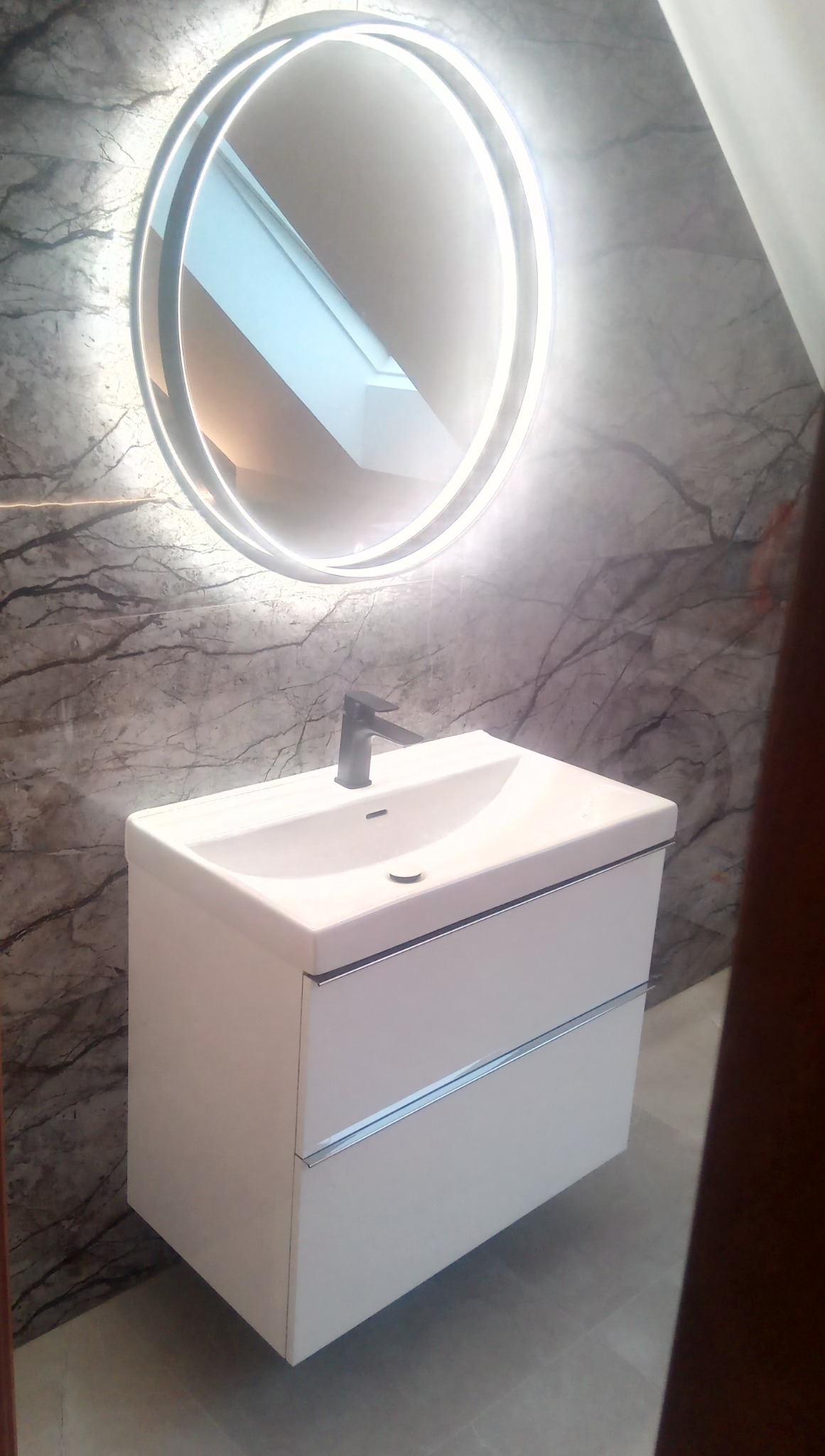 Biała szafka pod umywalkę z dwoma szufladami i czarną baterią, podświetlane okrągłe lustro na tle ściany z szarego marmuru.