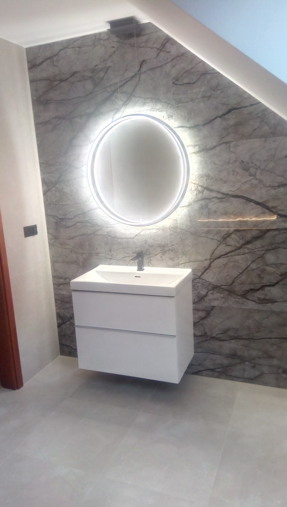 Nowoczesna łazienka z białą, podwieszaną szafką umywalkową i okrągłym lustrem z podświetleniem LED na ścianie wyłożonej płytkami imitującymi marmur.