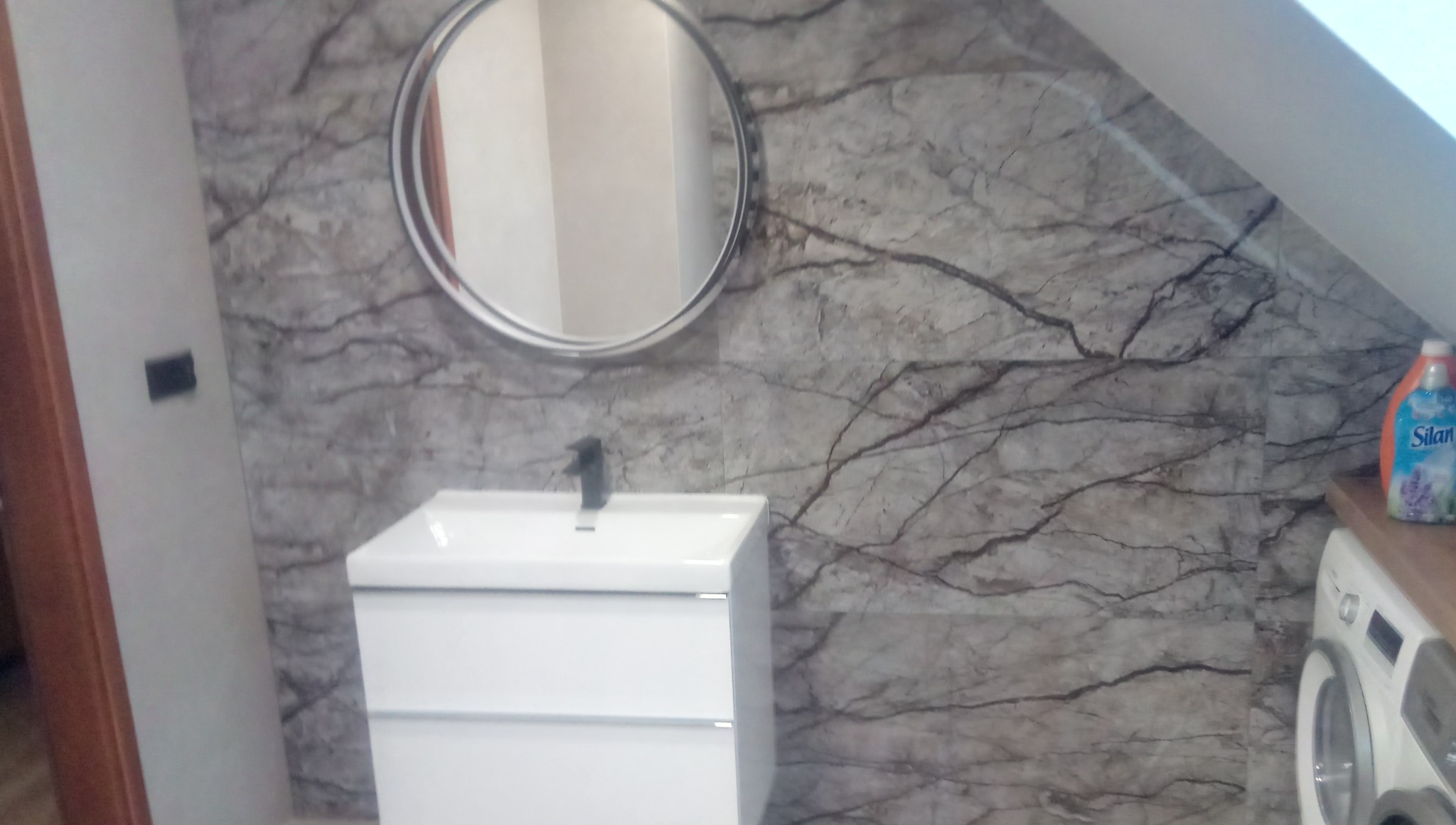 Wykończona łazienka na poddaszu ze ścianami z płytek imitujących marmur, okrągłym lustrem nad białą szafką z umywalką i pralką obok, uzupełniona butelką płynu do płukania.