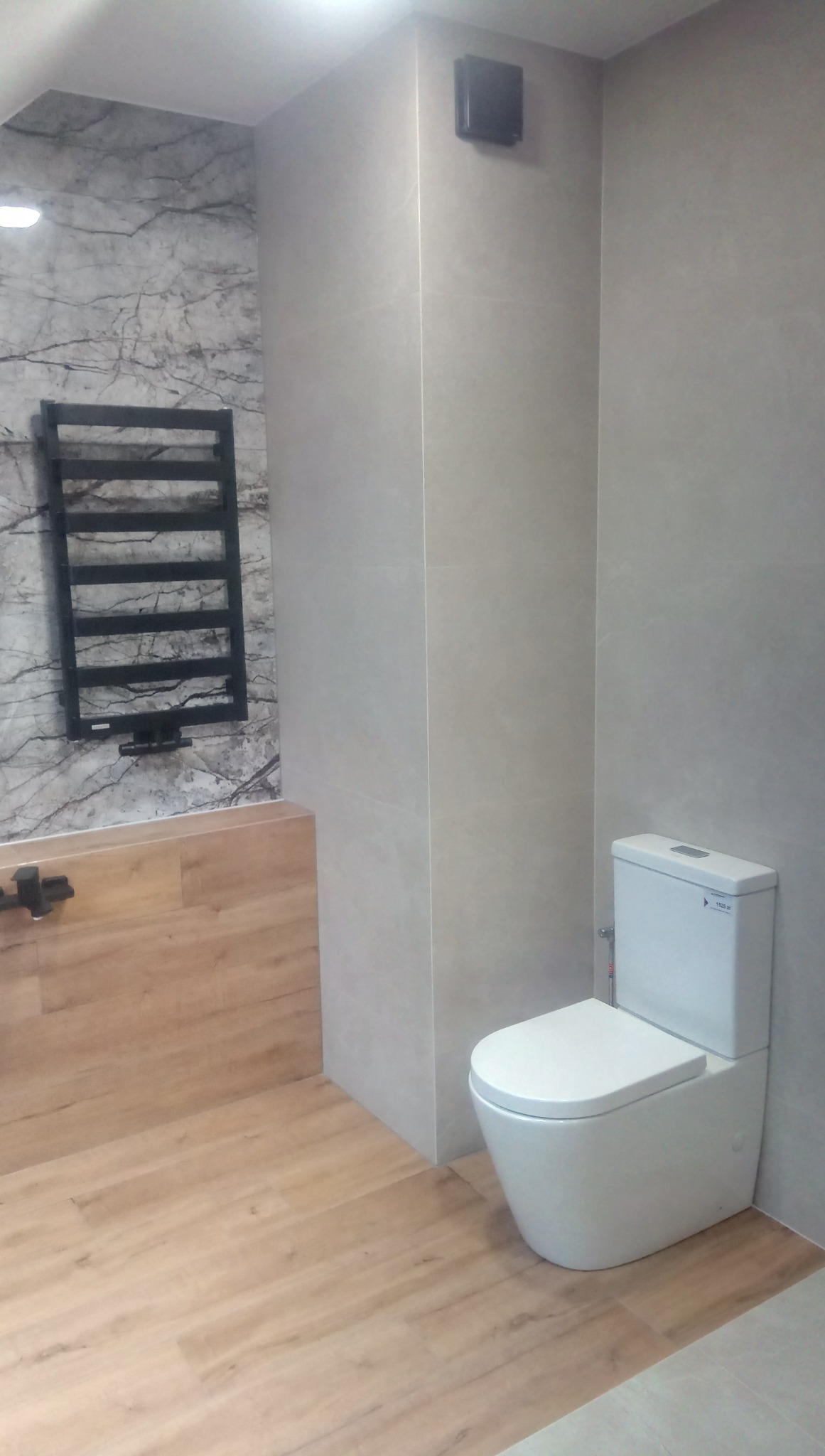 Nowoczesna łazienka z płytkami imitującymi marmur, drewnianą obudową wanny, grzejnikiem drabinkowym i białą toaletą kompaktową.