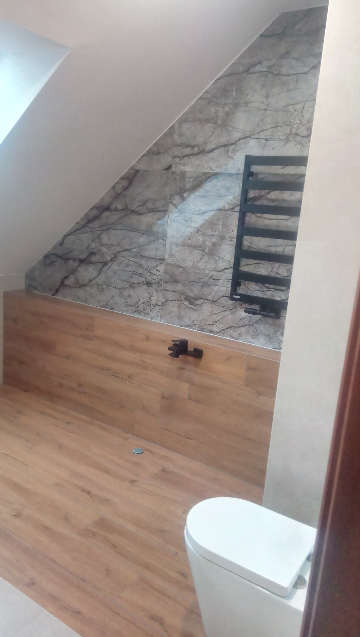 Ukończona łazienka na poddaszu ze skośnym sufitem, wykończona płytkami imitującymi marmur na ścianie, drewnopodobną okładziną na wannie i podłodze, z zamontowanym czarnym grzejnikiem drabinkowym...