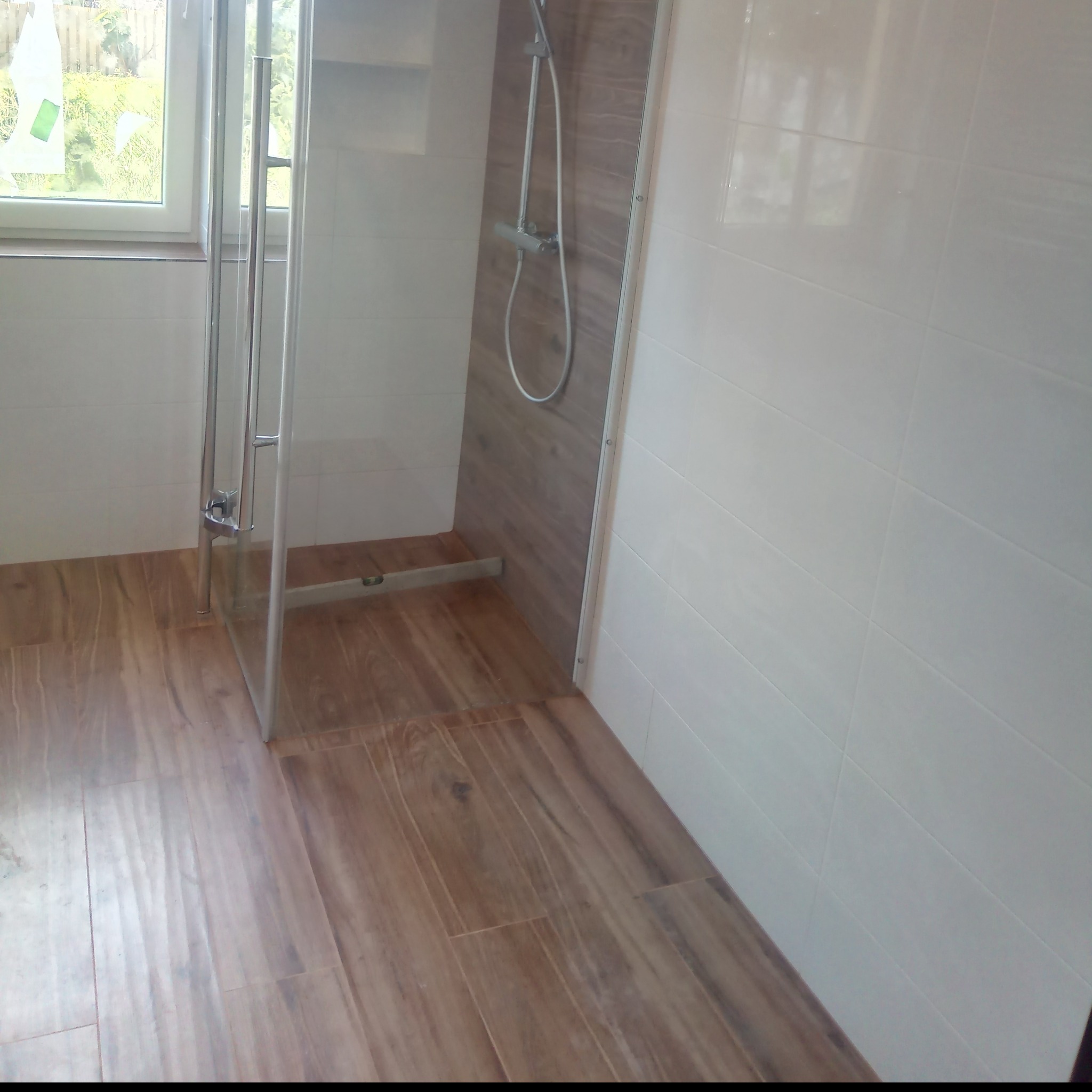 Nowoczesna kabina prysznicowa z przeszklonymi drzwiami, drewnopodobna podłoga i imitacja drewna na ścianie wewnątrz kabiny, białe płytki na ścianach łazienki.