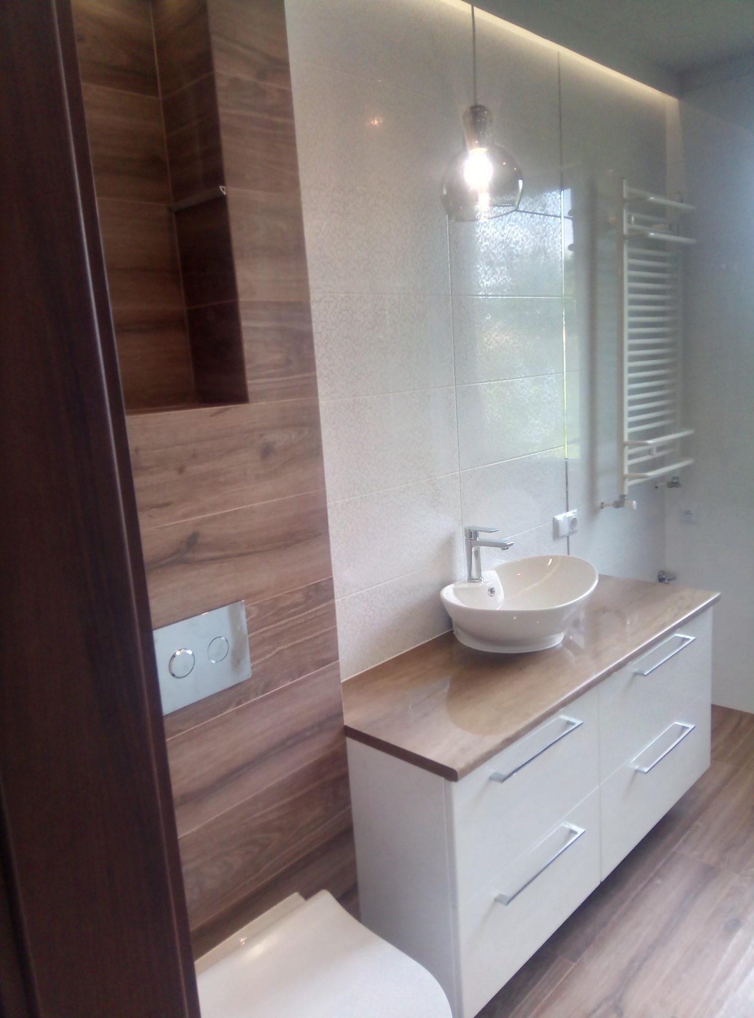 Nowoczesna łazienka z drewnopodobnymi płytkami, podświetlanym lustrem, umywalką nablatową na białej szafce z drewnianym blatem oraz grzejnikiem drabinkowym.