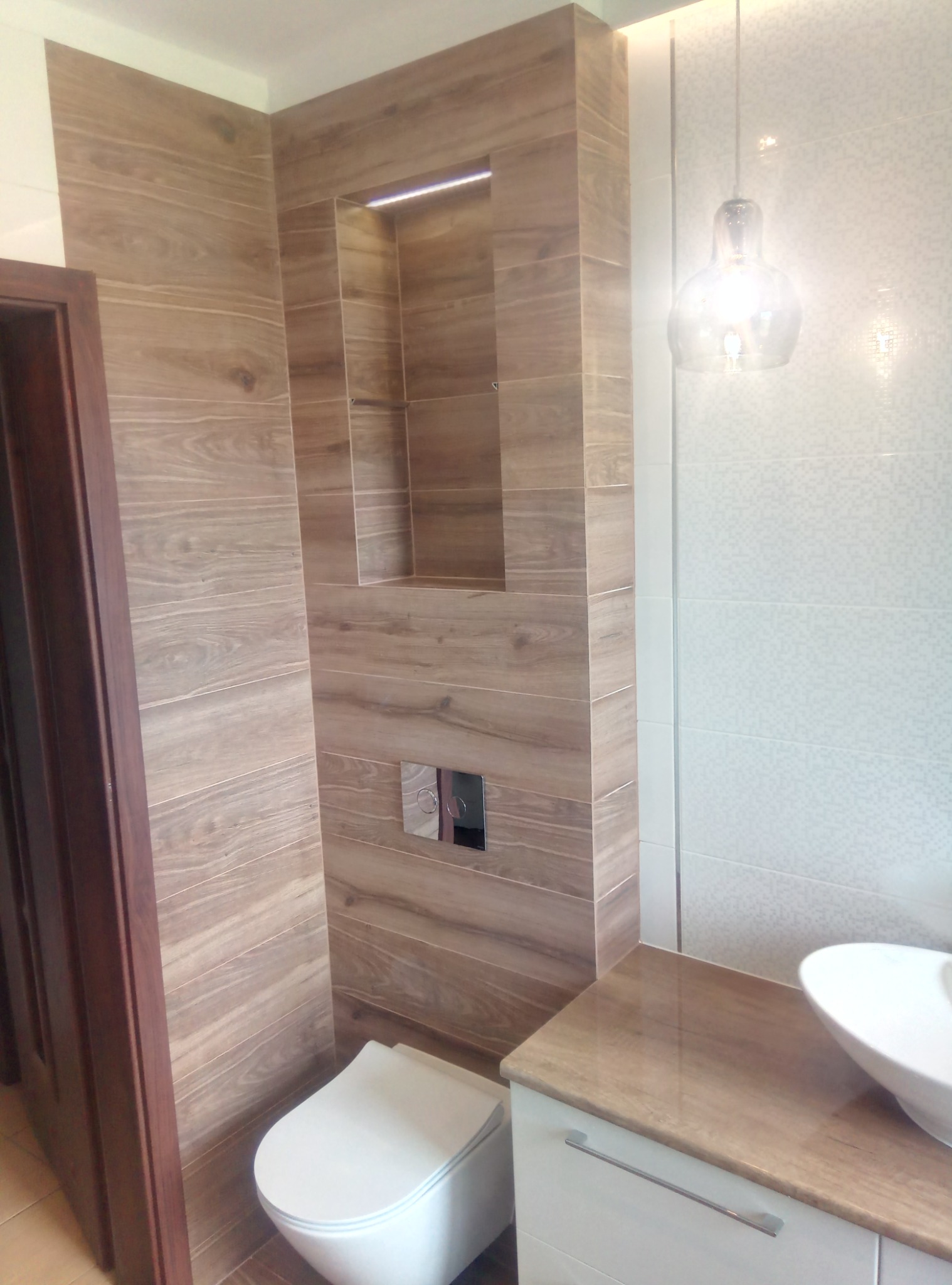Wykończona łazienka z drewnopodobnymi płytkami na ścianach, podświetlaną wnęką nad toaletą, wiszącą lampą i umywalką nablatową.
