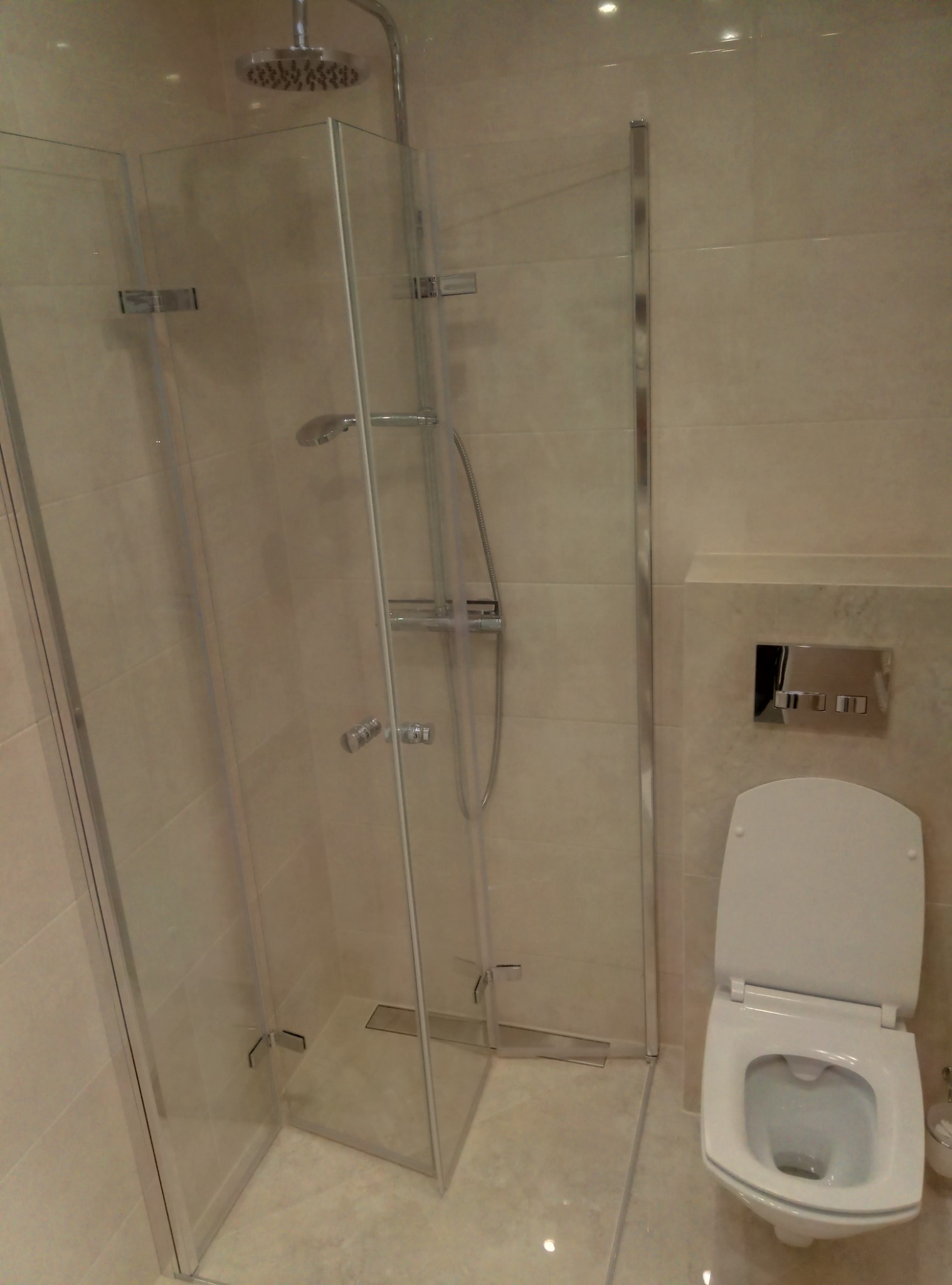 Nowoczesna kabina prysznicowa z deszczownicą i odpływem liniowym, obok toaleta podwieszana ze spłuczką w zabudowie, jasne płytki ceramiczne na ścianach i podłodze.