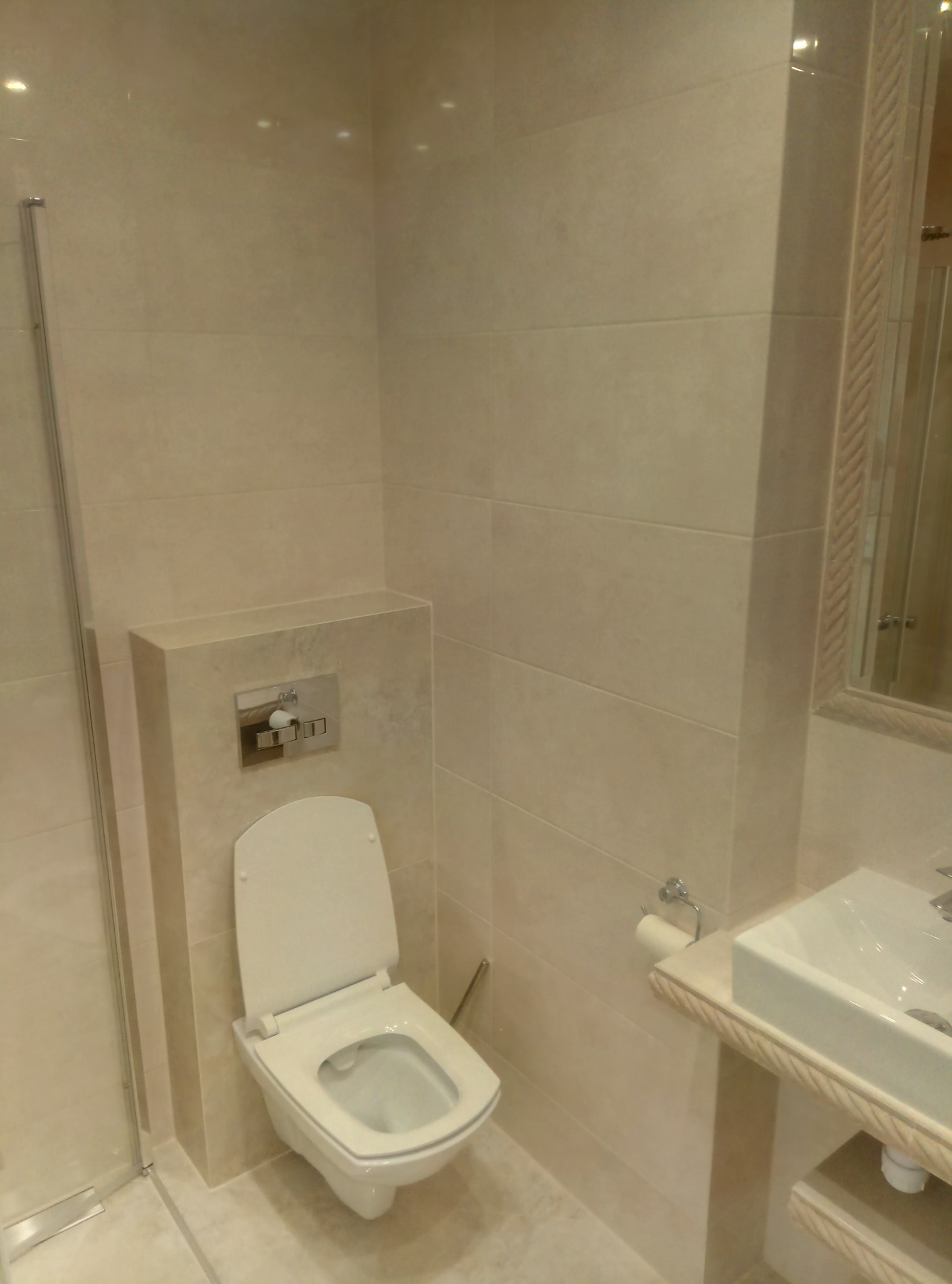 Nowoczesna toaleta podwieszana z zabudowaną spłuczką, wykończona beżowymi płytkami ceramicznymi, obok umywalka z lustrem w ozdobnej ramie.